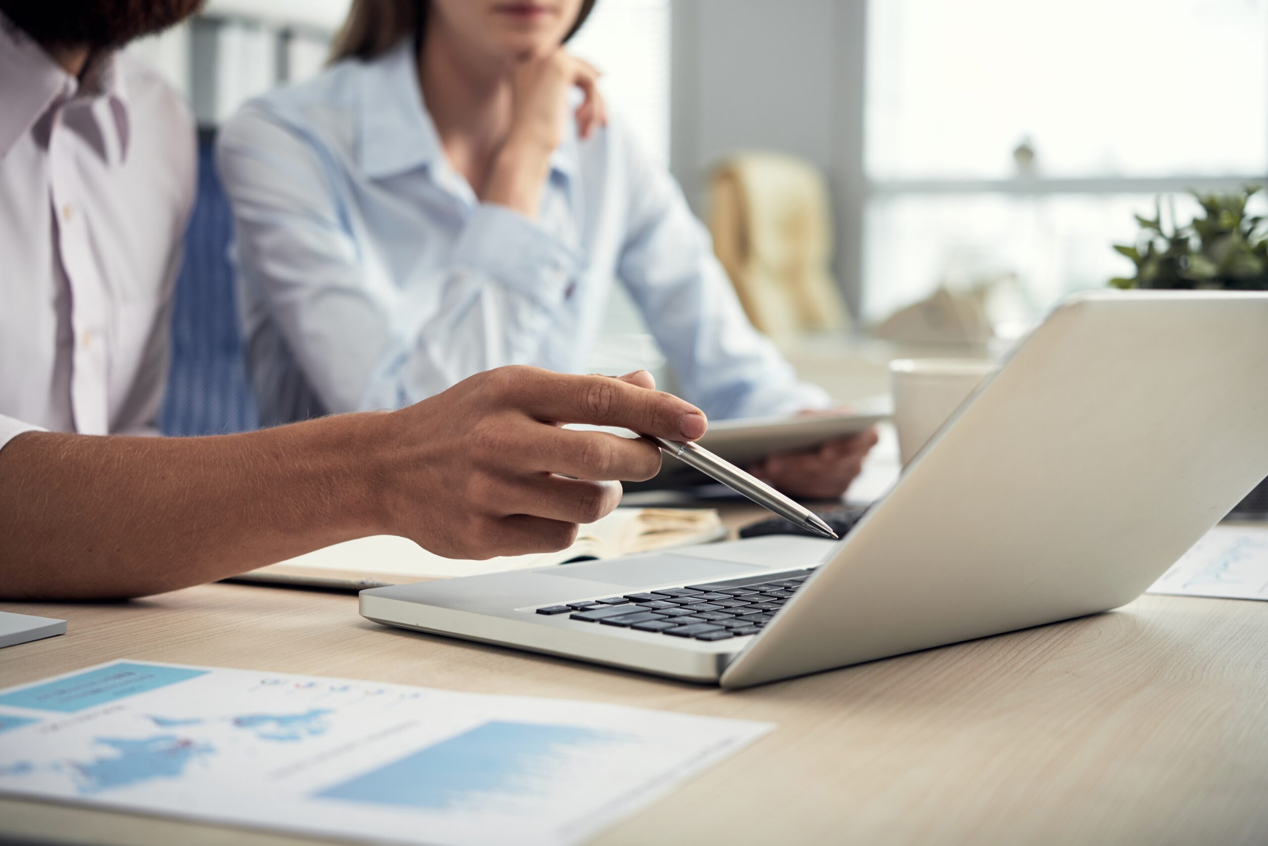 Two business professionals reviewing data on a laptop at a desk, with charts and documents visible, illustrating teamwork and digital collaboration.