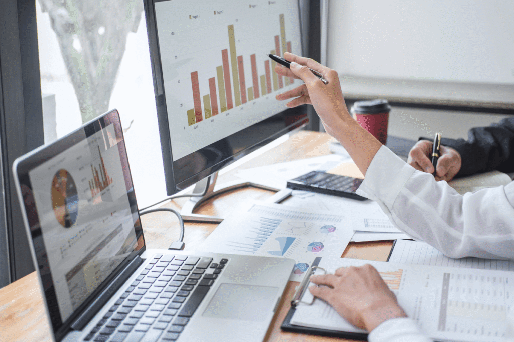 Business team reviewing performance charts on a monitor and laptop, surrounded by printed reports, a calculator and notepads.