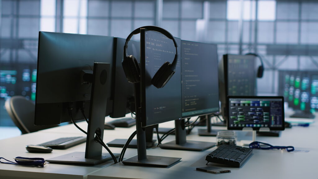 Multiple monitors and keyboards at an IT operations workstation, with headphones on a stand in a darkened technical environment.