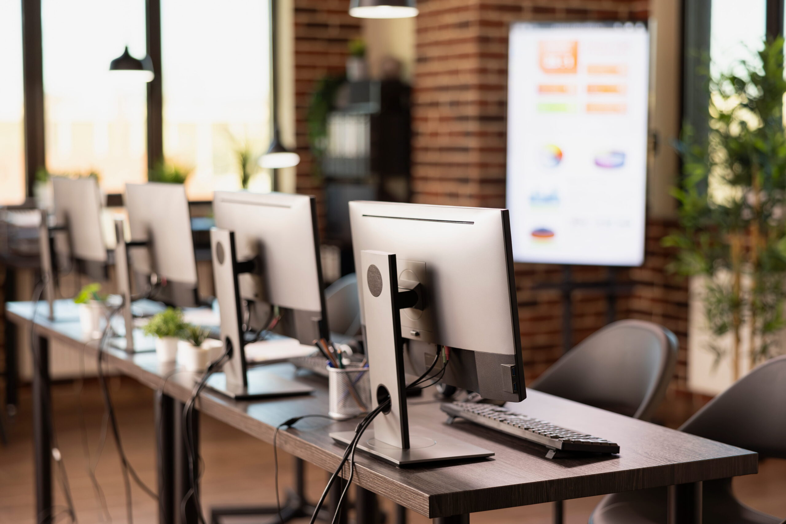 Row of desktop computers on desks in an empty modern office, with large windows and a presentation screen in the background.