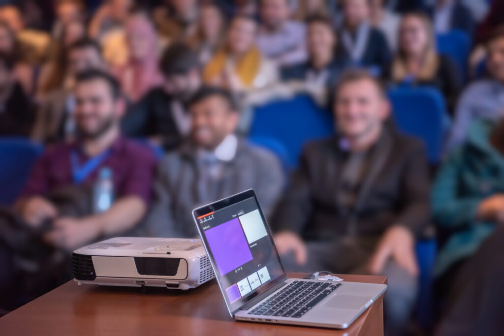 Laptop with a projector set up on a desk in front of a seated conference audience